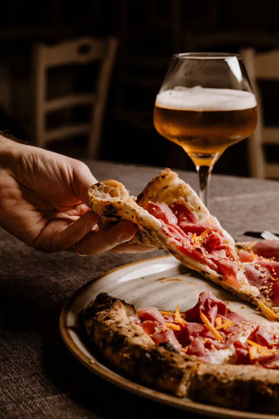 Trancio di pizza con crudo sollevato a mano davanti a un calice di birra